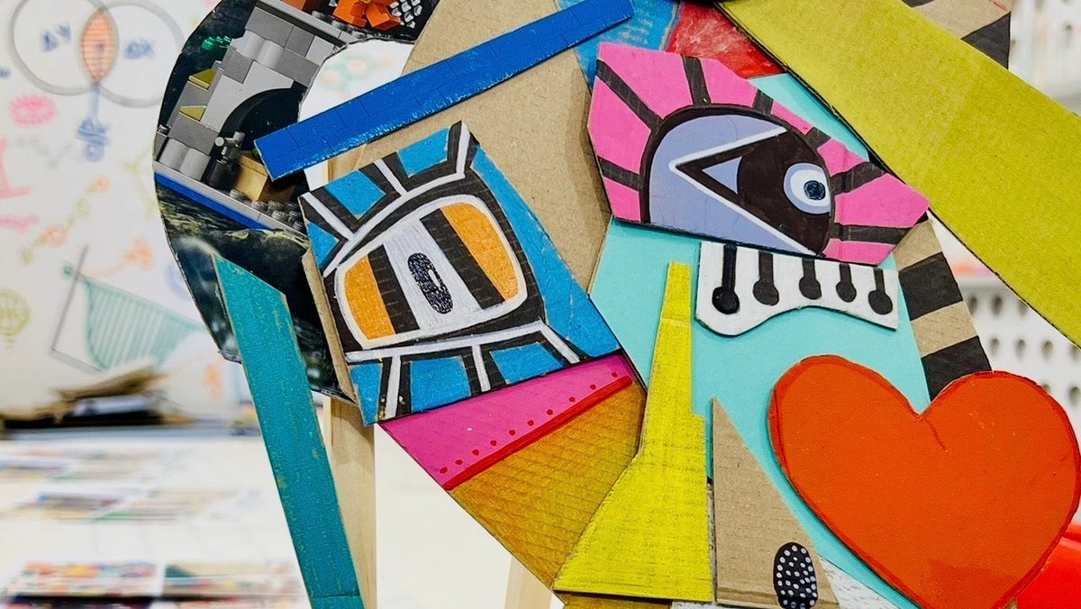 A colorful abstract art sculpture displayed on a wooden easel in a gallery or studio space. The piece features a patchwork of geometric shapes, including a large pink heart, a red heart, a yellow circle, a blue patterned triangle, and a black-and-white checkered triangle, with a central stylized face.